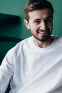 pexels photo 4065139 4065139 A happy man with a beard smiling while sitting indoors in a white t-shirt.