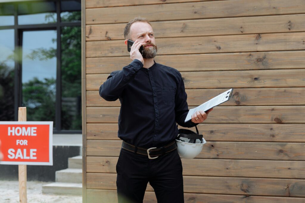 pexels photo 8482517 8482517 Real estate agent making a phone call while holding documents near home for sale sign.